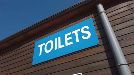 A clear blue sign indicating the location of public toilets, mounted on the wooden exterior of a modern restroom building