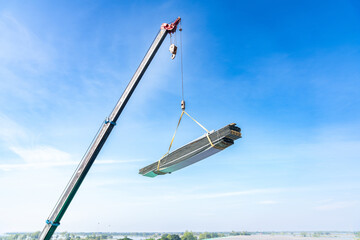 Mobile crane lifting a large bundle of metal or steel roofing sheets against a clear blue sky on a construction site.