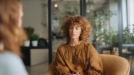 Young woman with curly hair attentively listens during a therapy session in a modern office. Natural light and greenery create a calm, supportive atmosphere for mental health conversations