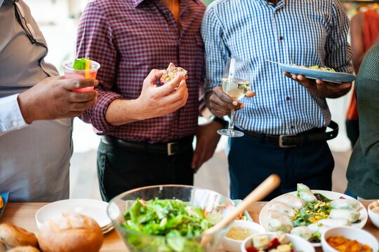 Group of people enjoying a meal, holding drinks, and chatting. Plates of food and salad on the table. Casual gathering with diverse individuals. Divers men gathering over food.