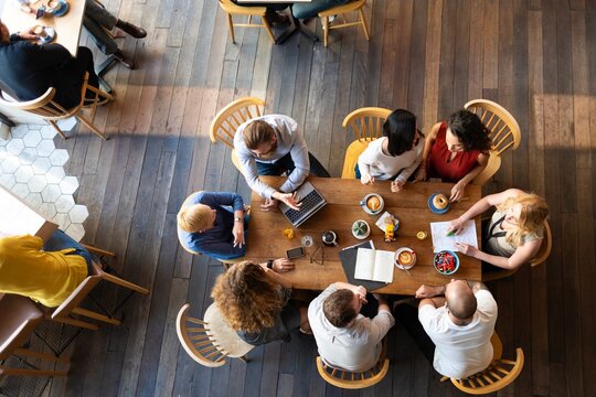 Top view of diverse group meeting at a table. People discussing, using laptops, and sharing ideas. Collaborative meeting with diverse team members. Casual startup business team working at a cafe.