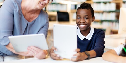 Smiling student boy uses a tablet in a library, guided by woman teacher. Diverse teacher and student hold tablets, engaging, learning activity together in the library. Teacher teaching boy in class