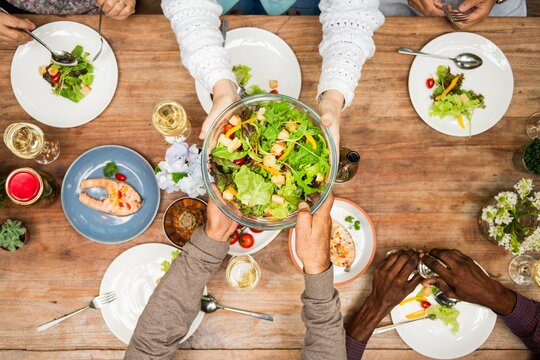Diverse group sharing fresh salad meal at wooden table. Hands passing salad bowl. Healthy dining, diverse gathering, sharing food. Top view of healthy food meal with diverse people. Healthy lifestyle