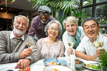Group of diverse elderly friends enjoying a meal together, smiling and holding wine glasses, celebrating friendship and happiness in a cozy setting. Happy diverse retired people celebrating at lunch.