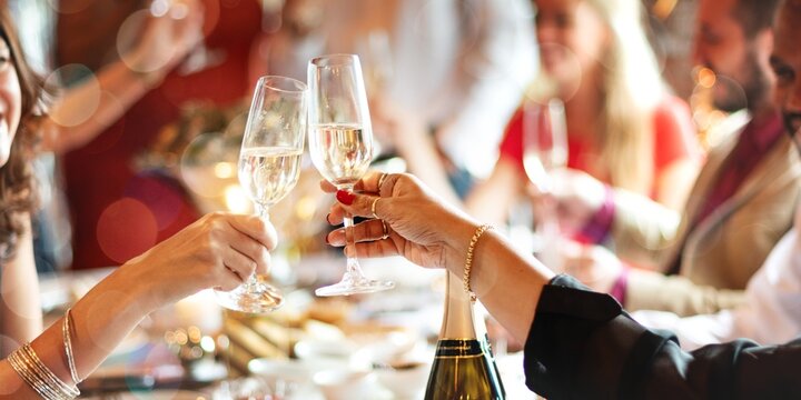 Two women toasting with champagne glasses at celebration party. Hands with champagne glasses celebrating at a festive dinner party, toasting with champagne surrounded by diverse group. - Powered by Adobe
