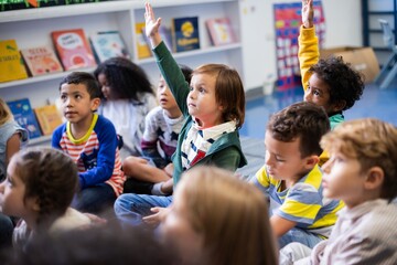 Children in a classroom sitting on the floor, raising hands. Diverse group of kids engaged in learning. Classroom setting with books and eager students. Little diverse kids raising hands in classroom.