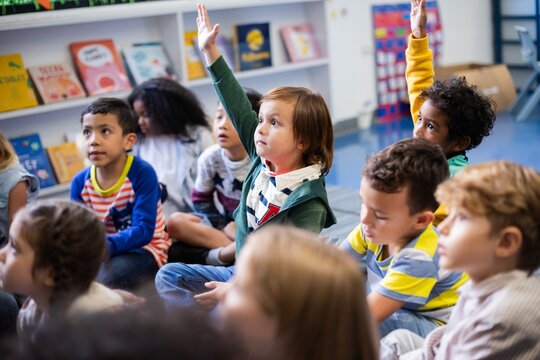 Children in a classroom sitting on the floor, raising hands. Diverse group of kids engaged in learning. Classroom setting with books and eager students. Little diverse kids raising hands in classroom. - Powered by Adobe
