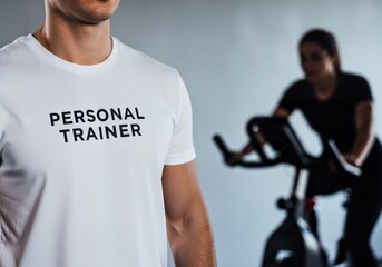 Male Personal Trainer Wearing White T-Shirt with Text While Woman Exercises in Gym