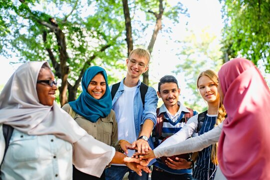 Diverse group of young adults in a park, smiling and stacking hands. Multicultural friends, teamwork, and unity. Outdoor gathering, friendship, and diversity. Diverse friends stacking hands in unity.