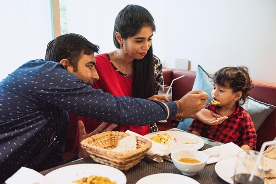 Indian family dining together, parents feeding child. Indian family enjoying meal, sharing food. Parents and child at table, bonding over dinner. Family time, sharing moments. happy Indian family.