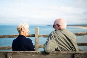 Elderly Caucasian couple enjoying a scenic sea view. Happy elderly couple sitting on a bench. Elderly couple sitting on a bench by the sea talking. Senior white man and woman at a bench by the sea.