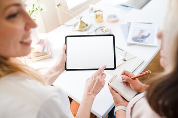 Two people collaborate at a desk, one holding a tablet, the other taking notes. Bright workspace, teamwork, and technology in focus. Fashion designers working together with tablet.