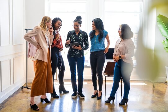 A diverse group of women in a bright office, discussing work. Diverse women standing together, smiling, and using a tablet. Professional and collaborative diverse group of women.