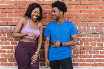 Two African American people smiling and standing against a brick wall, wearing casual sportwear. African American enjoying exercise. Happy African American couple wearing sportwear outdoor.