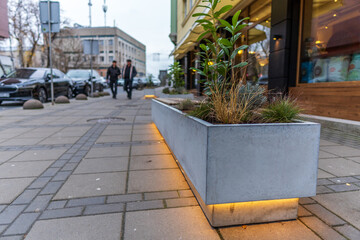 A modern concrete planter with lush green plants stands on a paved city sidewalk, softly illuminated from below, enhancing the urban street view.