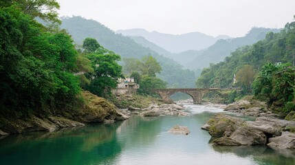 Scenic vista of a stone bridge arching over a clear river with lush green mountains and trees under a misty sky