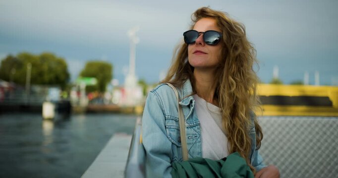 A Happy Young Woman with Blonde Curly Hair and Sunglasses Enjoys a Relaxing Ferry Ride on the Water, Looking at the City Skyline in Amsterdam, Netherlands, During Her Summer Travel Adventure.
