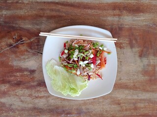 spicy Thai instant noodle salad served on a white plate, topped with chopped spring onions and accompanied by fresh lettuce, presented on a rustic wooden table.