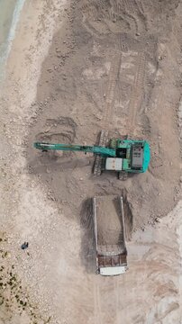 Aerial view of excavator reshaping sandy coastline, turquoise sea lapping, heavy machinery digging berms, shoreline,Vertical video