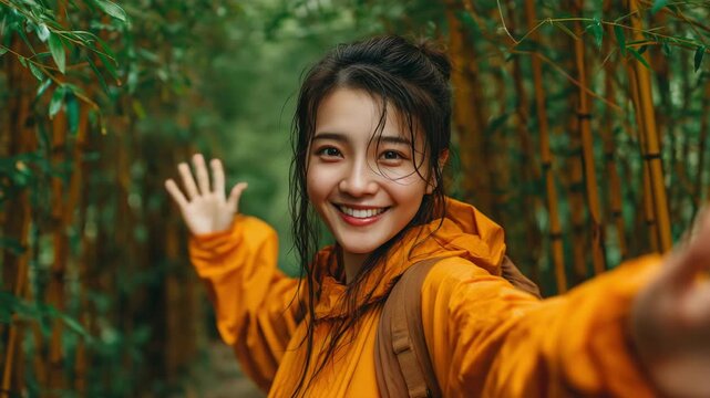 A joyful young woman wearing an orange jacket is seen in a photograph of a bamboo forest with a grin on her face and her hand outstretched toward the camera.
