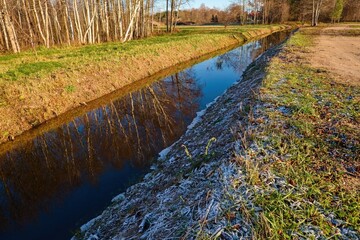 Frosty morning landscape with a calm canal reflecting bare trees, grassy banks, and soft sunlight in early winter.