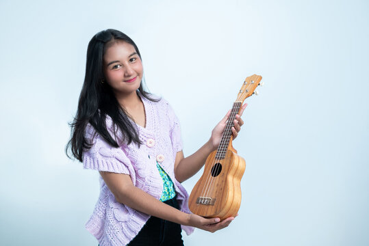 An Indonesian teenage girl holds a ukulele with a cheerful expression in a white studio background - Powered by Adobe