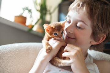 Young caucasian boy with brown hair hugging small orange abyssinian kitten. Child smiling gently, showcasing warm bond with pet. Sincere feelings. Copy space.