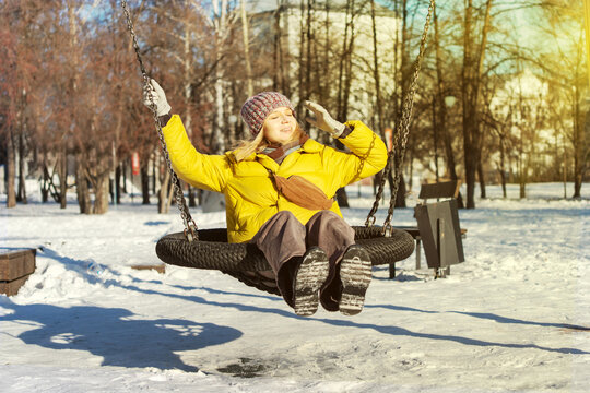Young woman in a yellow jacket enjoying a sunny winter day on a swing in a snowy park, smiling and feeling joyful in the bright sunlight. - Powered by Adobe