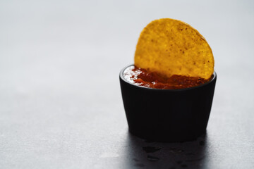 Round nachos in black ceramic bowl on concrete background with red salsa