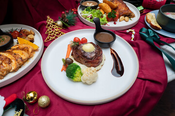 A delicious steak dish with vegetables, butter and gravy beautifully arranged on a plate surrounded by other dishes. The image is a festive scene, maybe Christmas. 