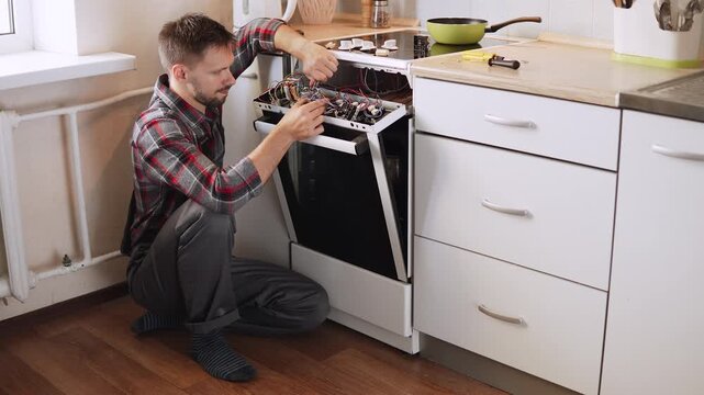 Caucasian man in overalls disconnects wires in disassembled electric stove control panel. Electrician repairs and maintains large appliances in kitchen.