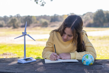 Girl researching sustainability concepts, writing in a notebook next to a miniature wind turbine and a globe, promoting environmental awareness and green energy education