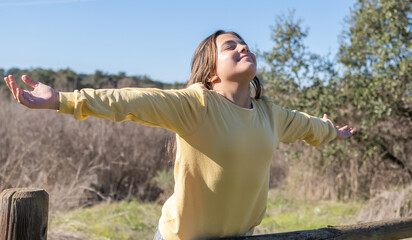 Young girl with arms outstretched, closing her eyes and feeling the sun on her face, experiencing happiness and well-being in a natural outdoor environment © Nanci