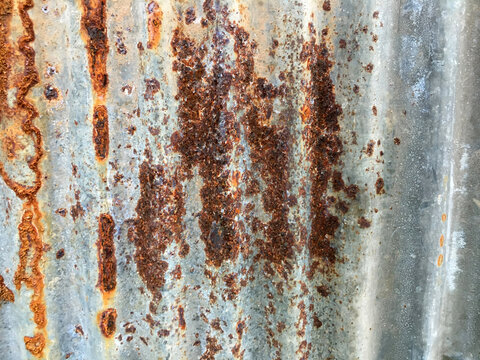 Full-frame view of heavily weathered and rusted corrugated metal sheeting, texture features distinct patches of vibrant orange, red, and deep brown rust against the faded, galvanized gray-blue surface - Powered by Adobe