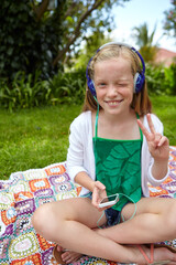 Portrait, child and headphones with phone at park for travel, streaming podcast and summer holiday. Smile, kid and peace sign for listening to music, download playlist and relax on grass for vacation