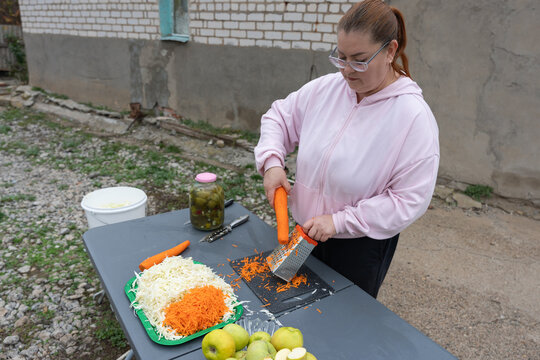 Rural woman shredding carrots and cabbage for winter pickling in her courtyard, preparing homemade sauerkraut and carrot salad on a plastic table with jars and apples nearby