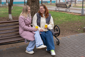 Two friends sitting on a park bench in autumn drinking coffee from yellow paper cups while laughing and chatting in casual coats and jeans, enjoying a cozy outdoor friendship moment