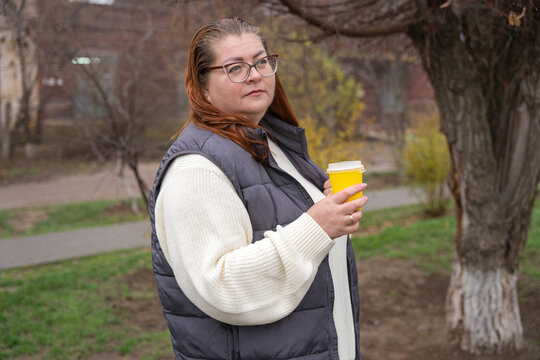 Comfortable plus size woman sips coffee from a yellow takeaway cup in an autumn park, wearing a cream knit sweater and dark puffer vest while strolling a leafy path with a relaxed happy smile