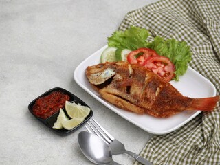 Fried Tilapia or Ikan Nila Goreng. Served on white plate with sambal (chilli sauce), cucumber and tomato slice. Selected focus.