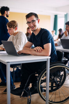 Portrait of physically disabled happy young man sitting with laptop at desk in high school classroom