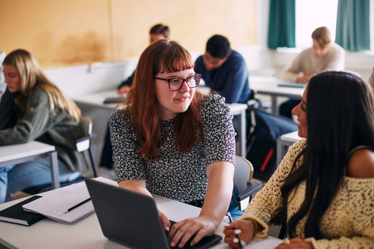Smiling teenage girl helping female classmate with studies on laptop while sitting at desk in high school classroom
