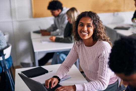 High angle portrait of smiling teenage female student sitting with laptop at desk in classroom at school