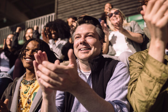 Smiling male spectator clapping while enjoying game in stadium stand at sunny day