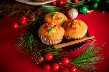 A warm and festive still life of Christmas-themed cupcakes topped with colorful candied fruit. The cupcakes are arranged with holiday decorations including pine branches, red berries, and rustic.
