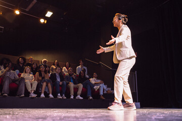 Full length of smiling female presenter wearing headset with arms outstretched talking in seminar at illuminated theater