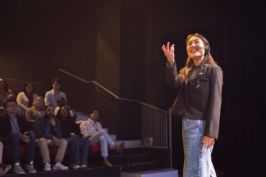 Smiling young female keynote speaker wearing headset gesturing while talking in seminar at theater