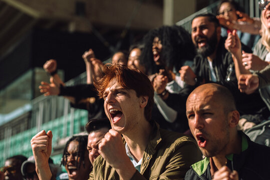 Excited young man shouting while cheering and making fist at sports stand on sunny day