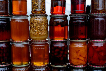 Colorful jars filled with fruit preserves, syrups, nuts, and honey are stacked together, showcasing homemade artisanal food products at a market.
