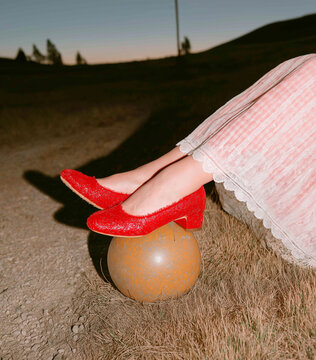 A woman in a pink velvet dress poses with sparkling red shoes balanced on a rusty ball in a field at dusk. Whimsical fashion scene with a rustic twist.