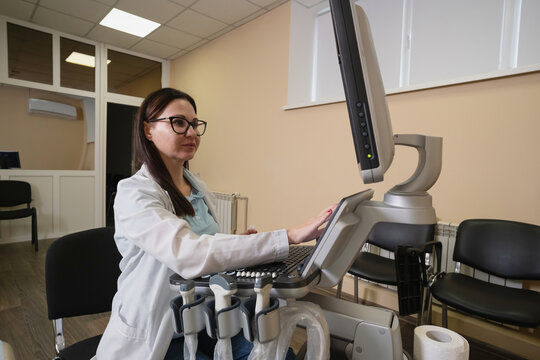 Female medical professional in a white lab coat and glasses operating an ultrasound machine. Performing an examination in a clinic setting. Focusing on healthcare technology and diagnostic procedures